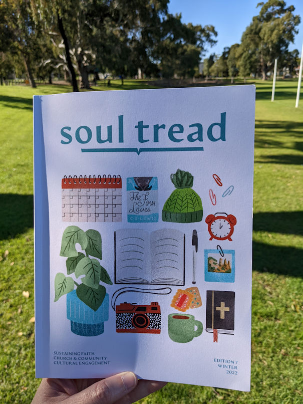 Photo of the cover of Soul Tread Edition 7 being held up in front of grass, trees and blue sky in the background.